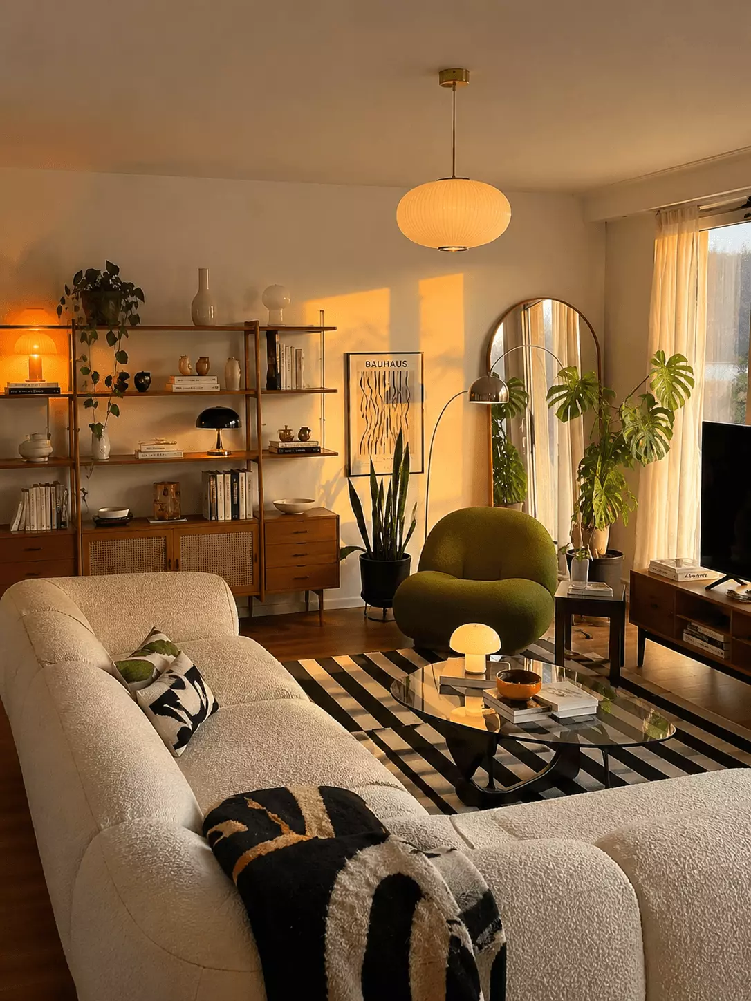 Bauhaus color mid-century modern living room with a cream modular sofa, olive accent chair, glass coffee table, cane bookshelf, wood TV console, indoor plants, paper pendant lamp, and black-and-white striped rug.