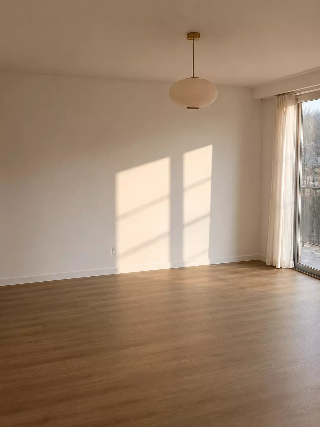 Before photo of an empty living room with white walls, wood flooring, a window, sheer curtains, and a pendant light before a mid-century modern redesign.