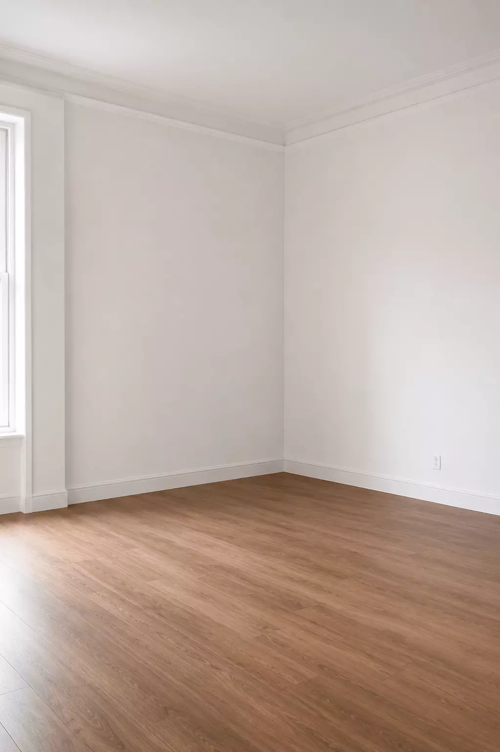 Before photo of an empty white living room with wood flooring, plain walls, crown molding, and a window before a mid-century modern redesign.