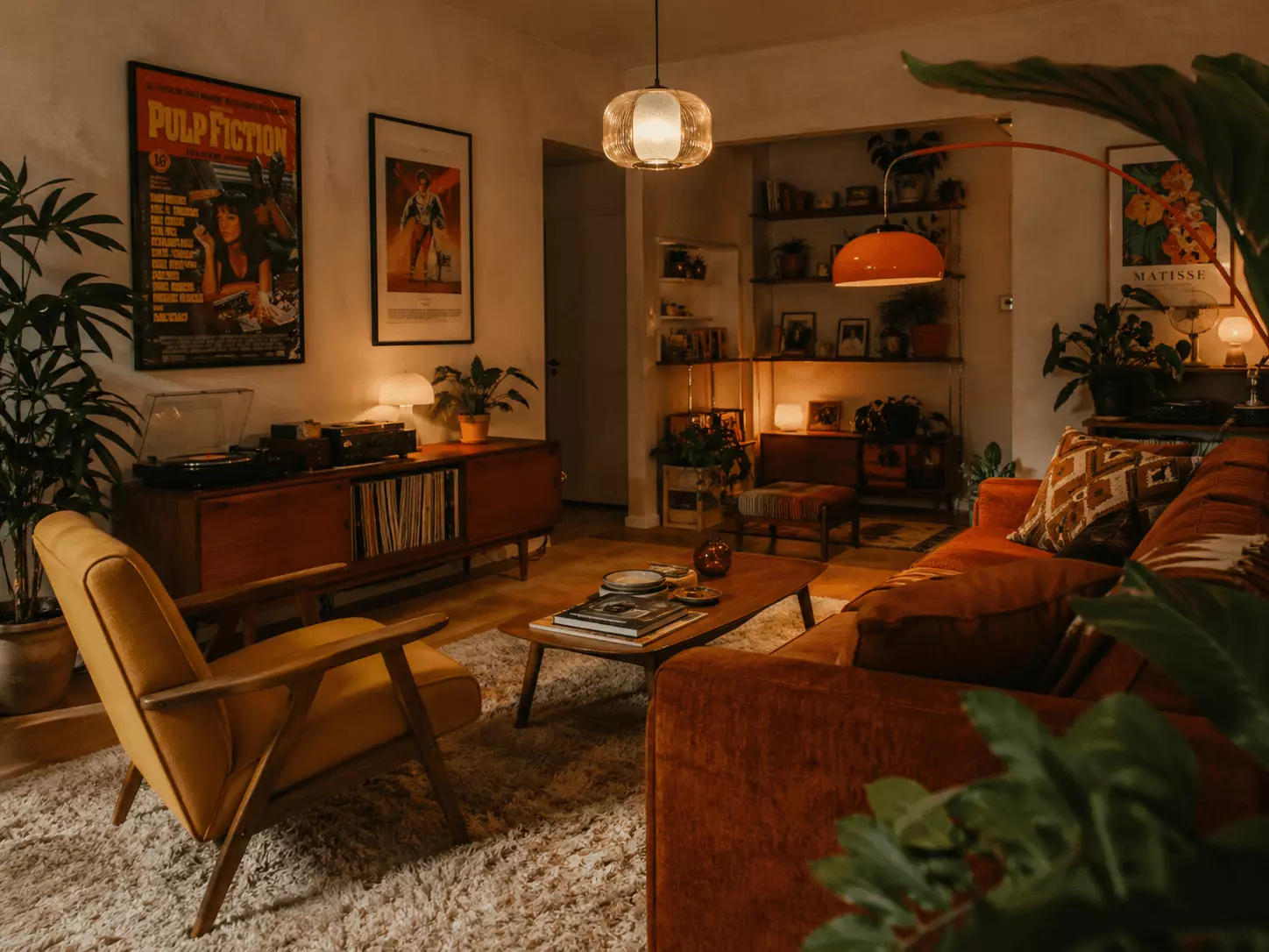 Orange retro mid-century modern living room with a burnt orange sofa, mustard lounge chair, wood coffee table, record player, retro posters, indoor plants, and warm lighting.